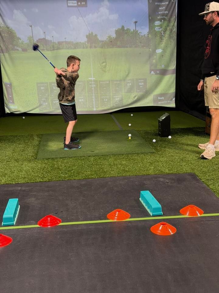 Boy swinging golf club indoors, instructor watches.  Golf simulator screen visible, practice setup with cones.