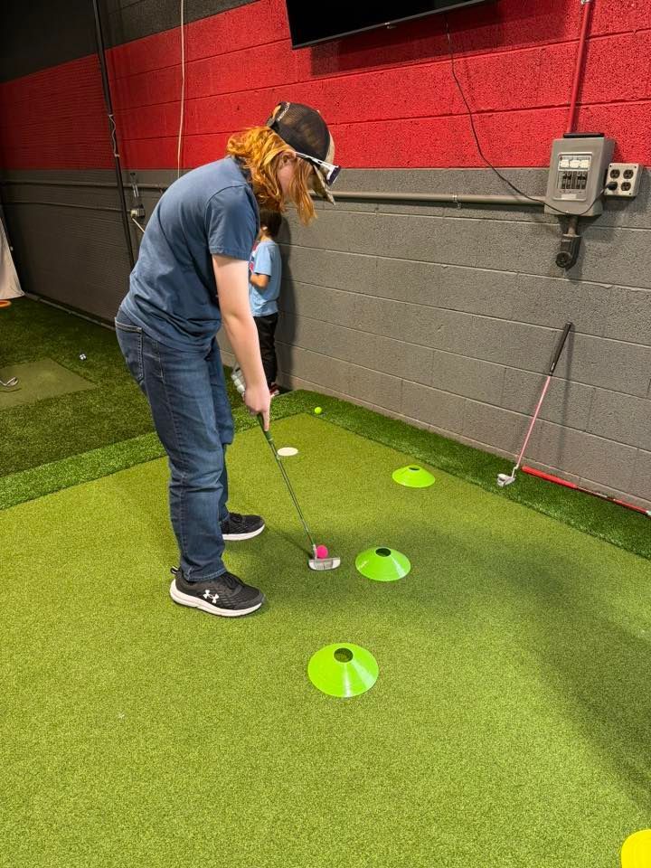 Person putting a golf ball on a mini-golf course. They wear a cap and blue shirt, and the green is marked with cones.