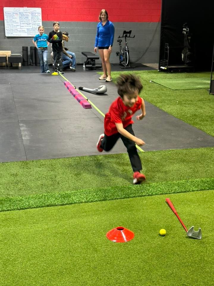 A child in a red shirt runs towards a miniature golf hole on turf; others observe in a gym.