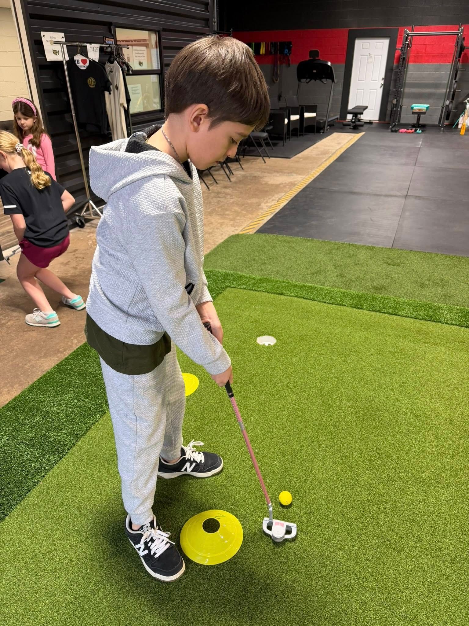 Boy putting on indoor putting green, wearing grey sweatsuit and black sneakers. Yellow cone marker, golf ball, and putter visible.