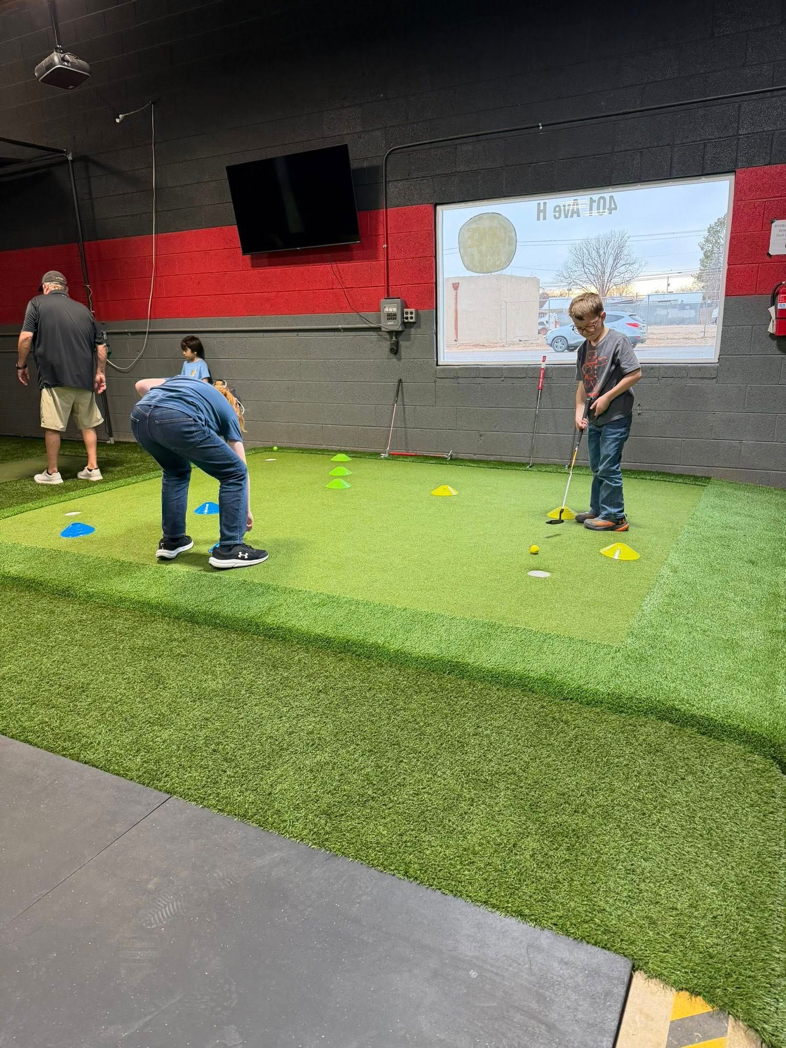 People playing mini golf indoors on green turf. One person putts. Cones and a map are visible.