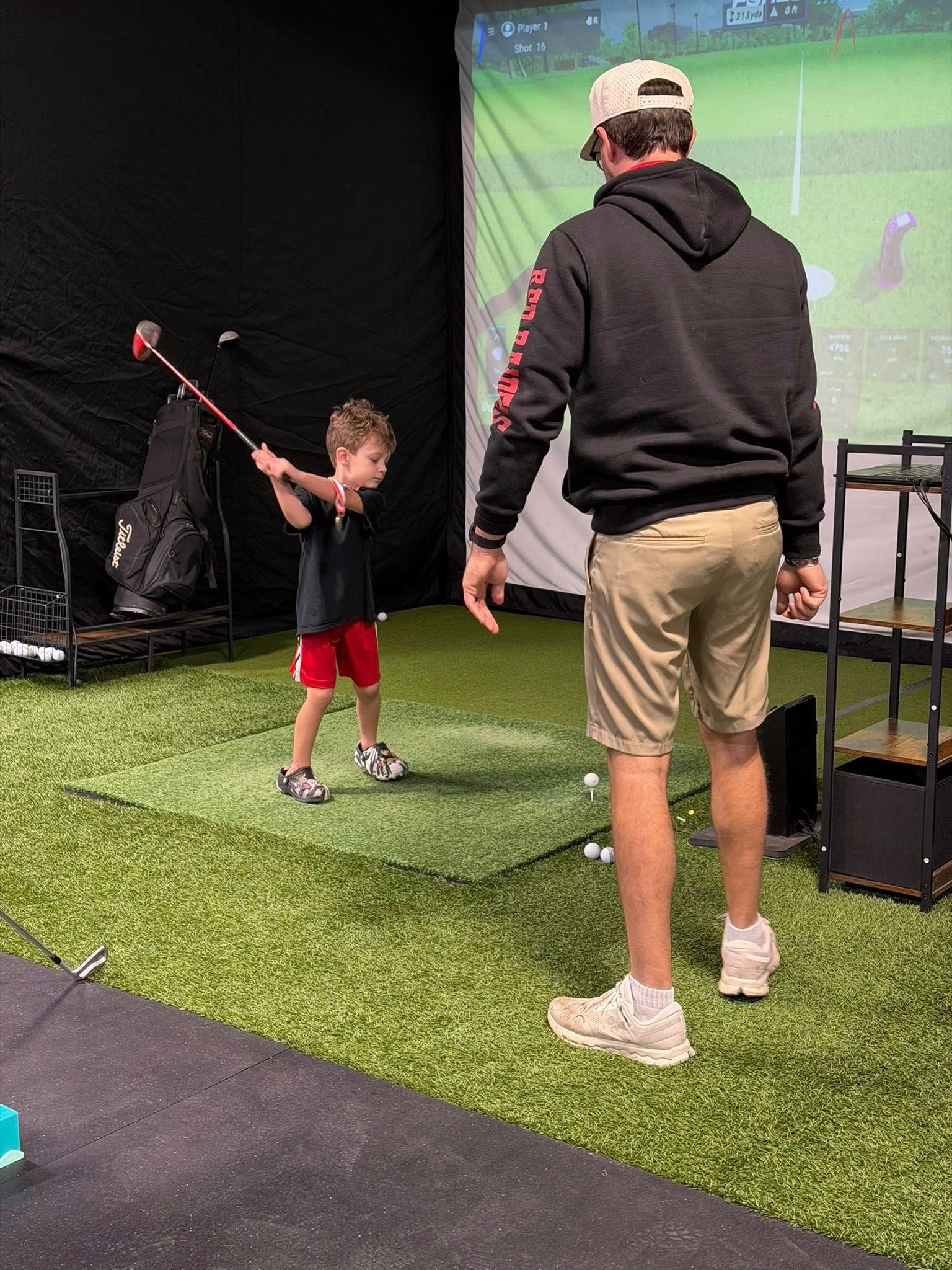 Young child swinging a golf club, with an adult watching on a green turf. Indoor setting.
