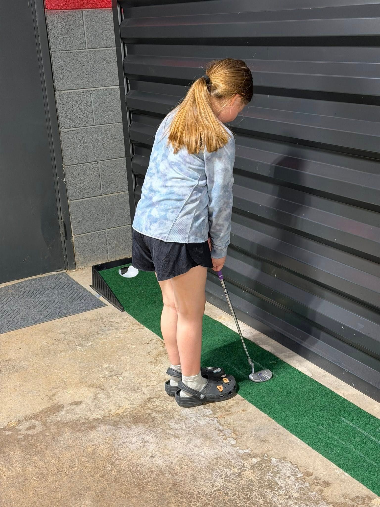 Girl putting a golf ball on a mini-golf course. Wearing a light blue jacket and black shorts.