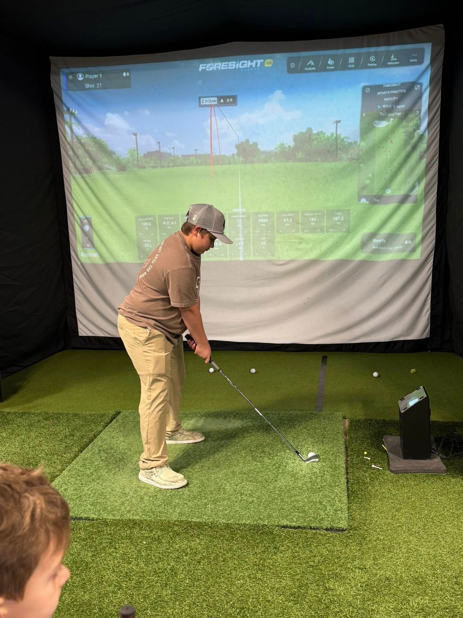 Person swinging a golf club in a golf simulator. He wears a hat and khaki pants.