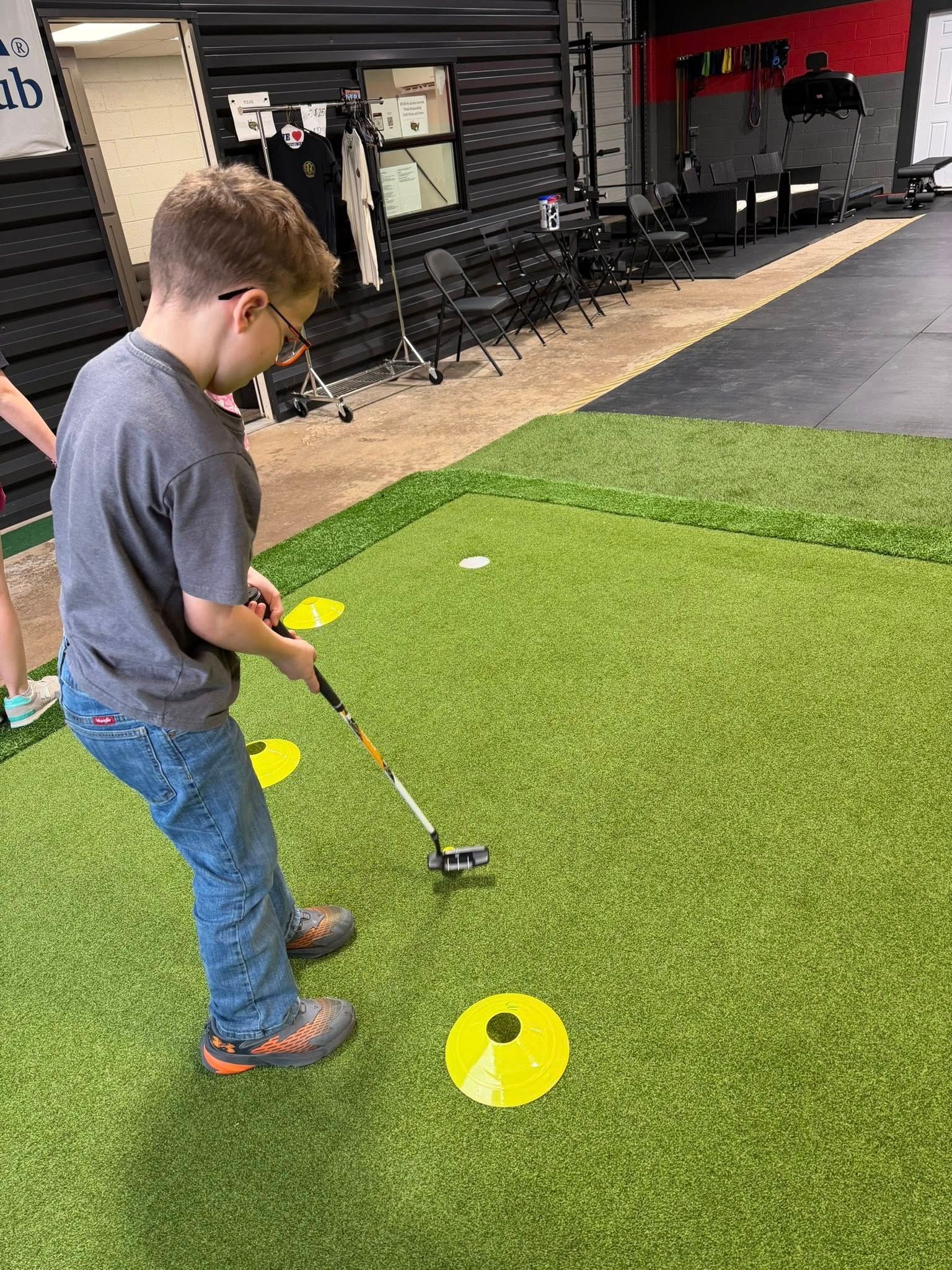 Boy with glasses putting on a mini golf course indoors, wearing jeans and a gray shirt.
