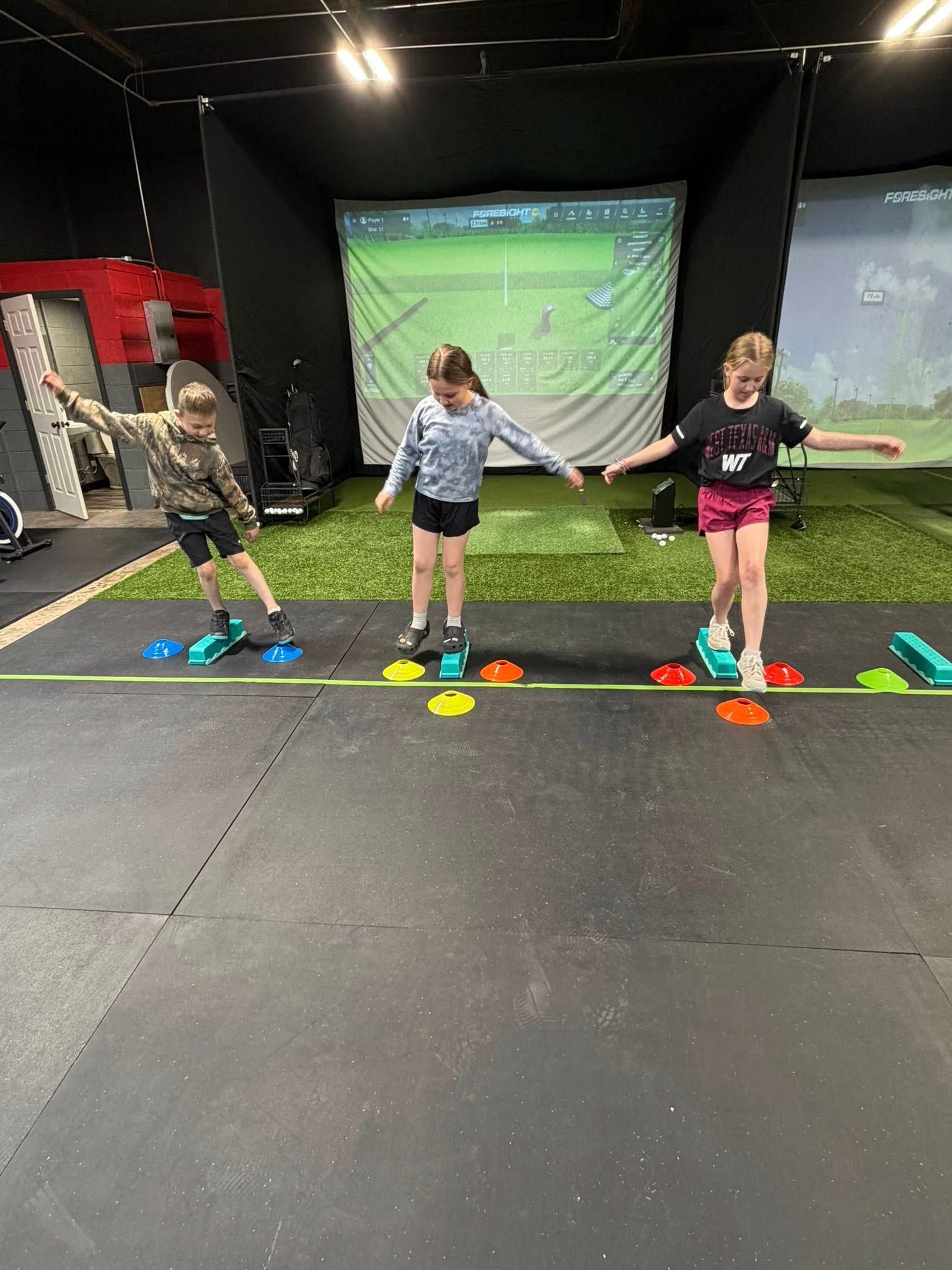 Three children balance on stepping stones in a gym with golf simulators in the background.