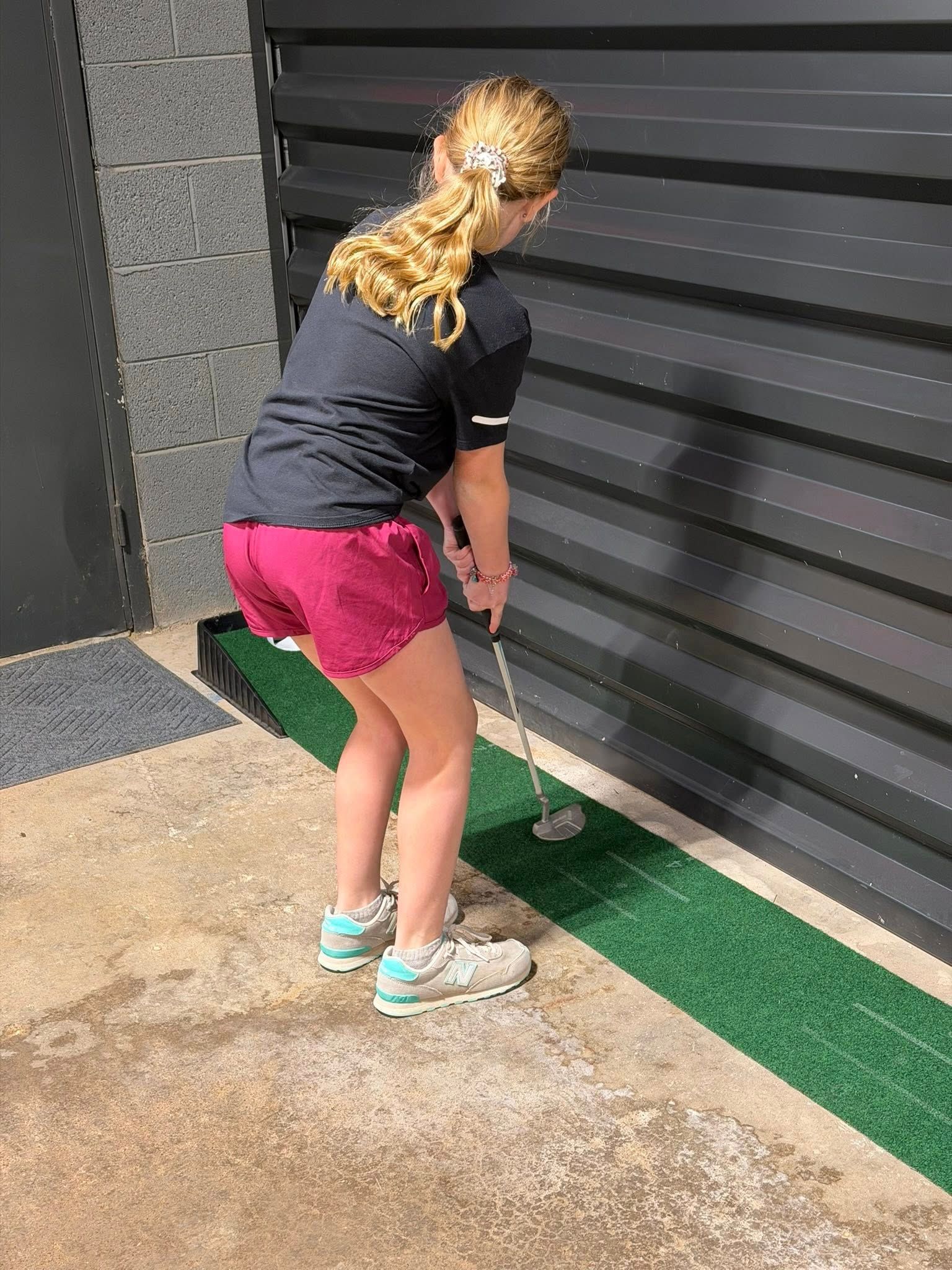 Girl putting a golf ball on a green mat against a black wall.
