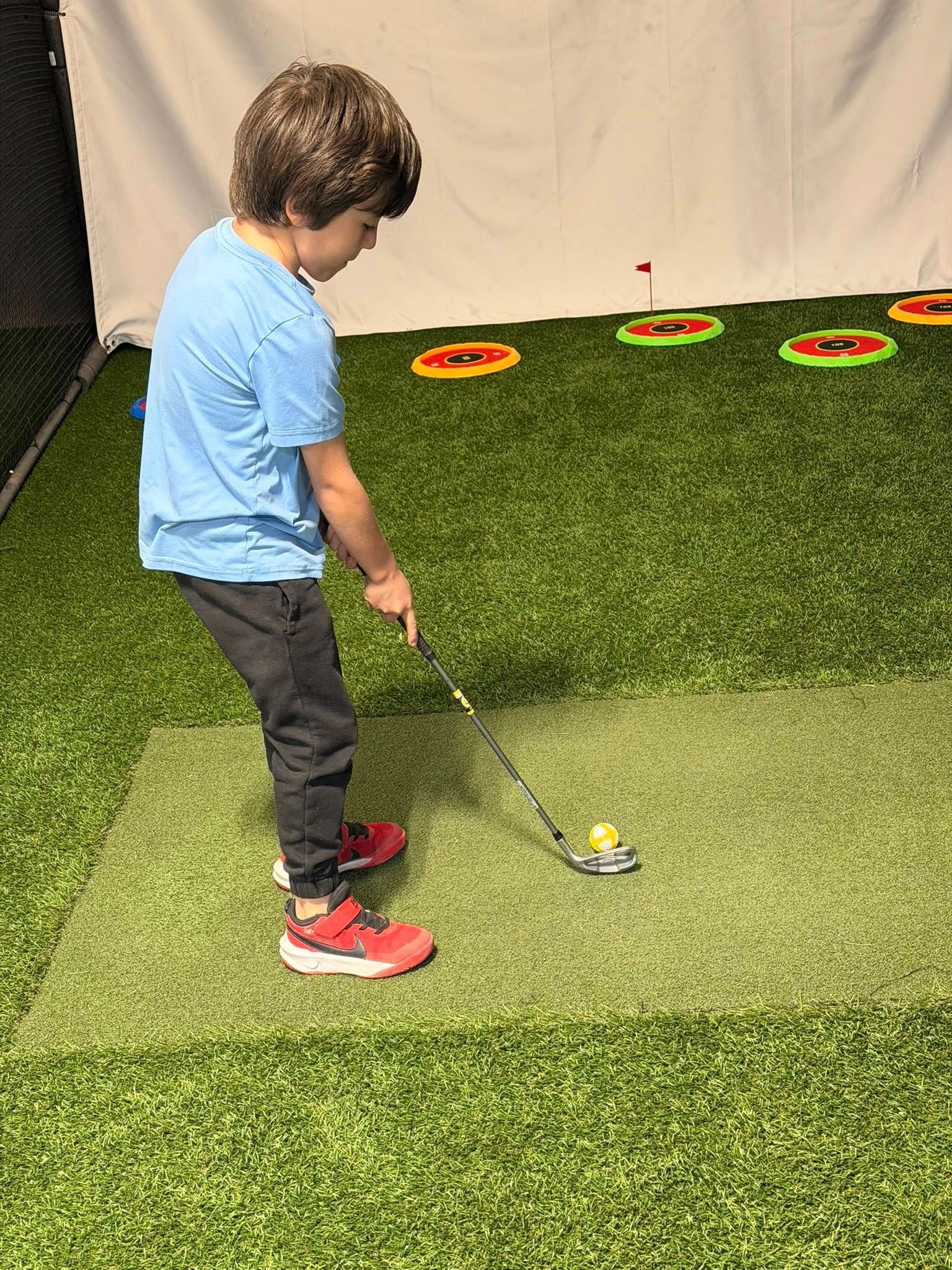 A child in a blue shirt and black pants is practicing golf, swinging a club at a ball on green turf.
