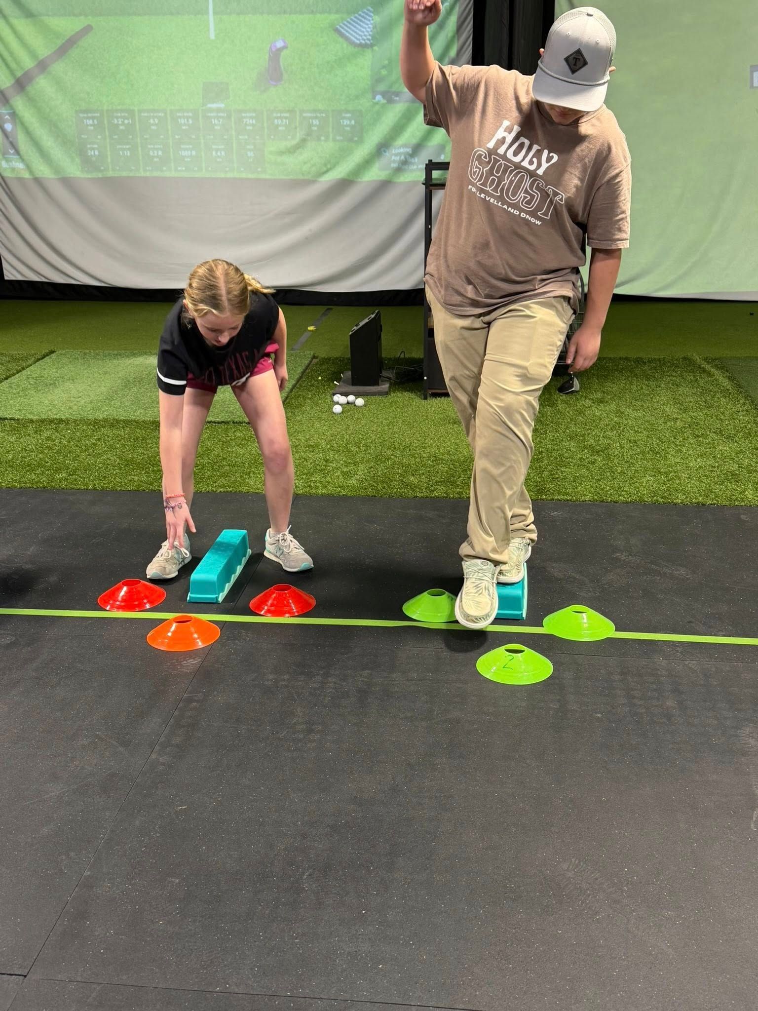 Two people in a gym setting are doing exercises with cones and blocks. One raises their arm.
