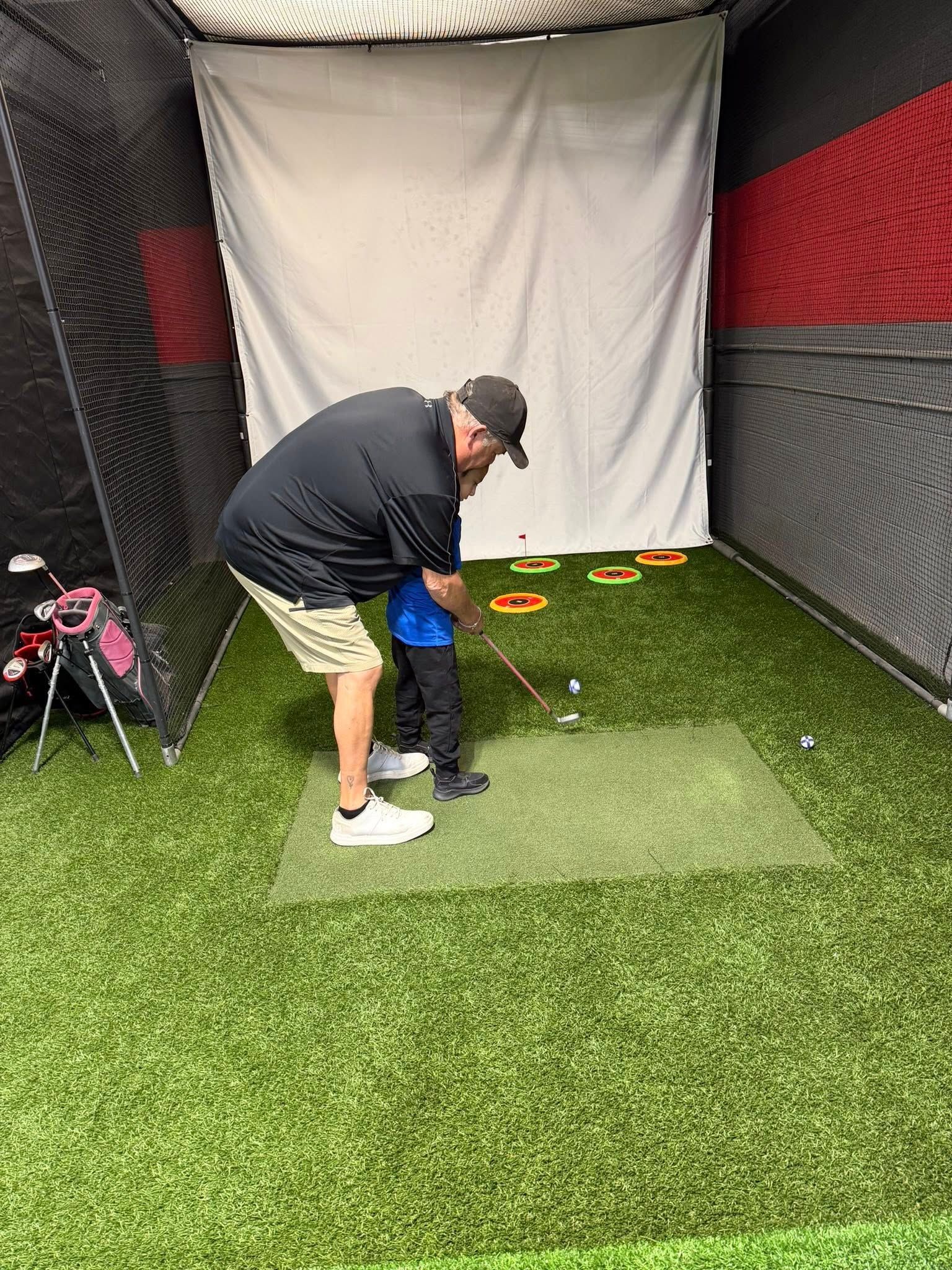 Man teaching a child to golf in an indoor golf simulator. They are on green turf; the child is holding a club.