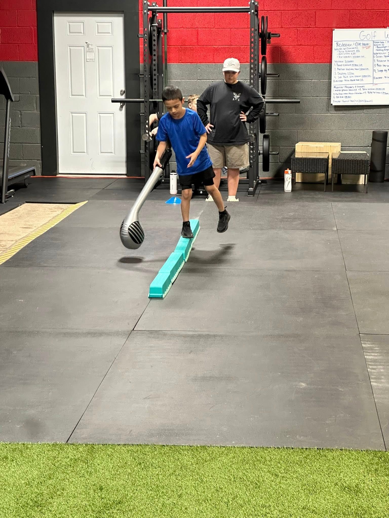 A boy balances on a blue beam, holding a weighted stick, with a coach observing in a gym.