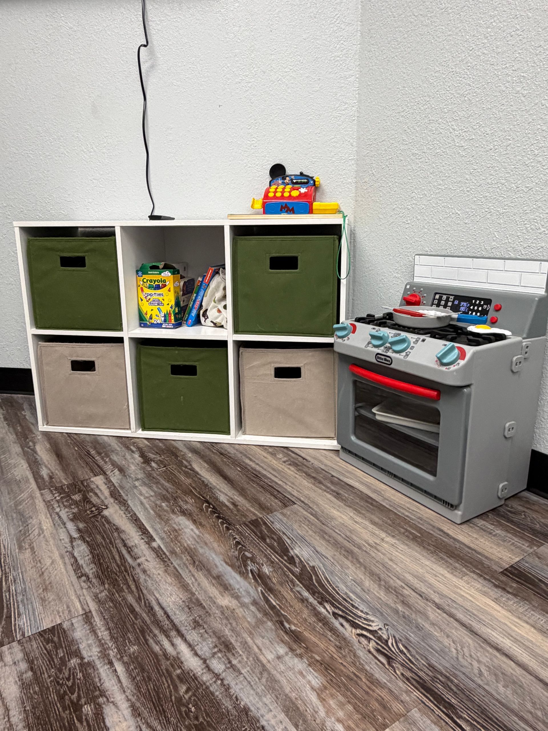 A child's play area with a white cube shelf, storage bins, and a toy kitchen set on a wood-look floor.