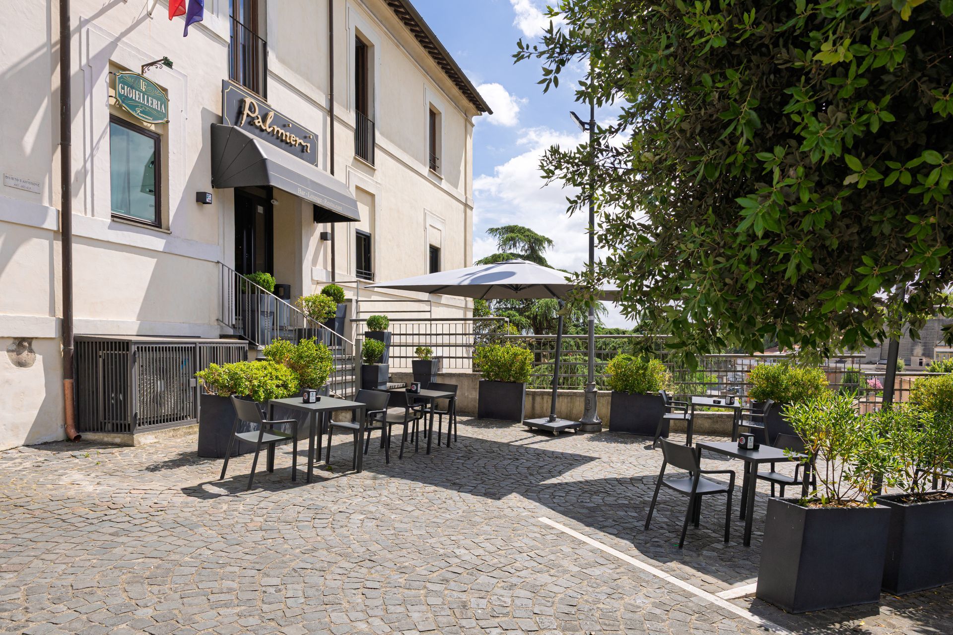 Dehor esterno del caffè con tavoli e sedie neri accanto a un edificio beige, sotto la luce del sole