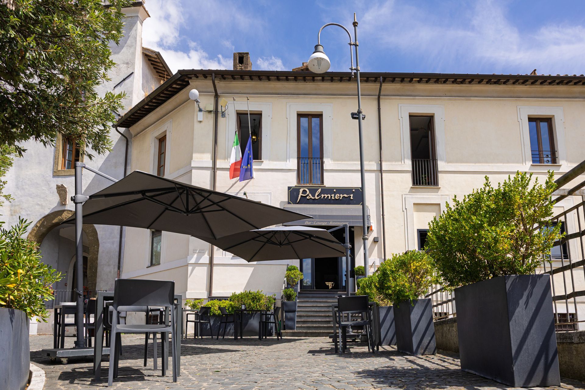 Terrazza esterna del caffè con ombrelloni neri e sedie di fronte a un edificio color crema con bandiere italiane.