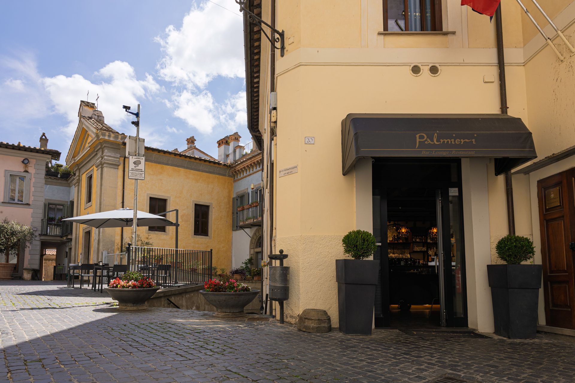 Una soleggiata strada acciottolata con edifici gialli, tende da sole dei caffè e una fioriera in una piazza europea.