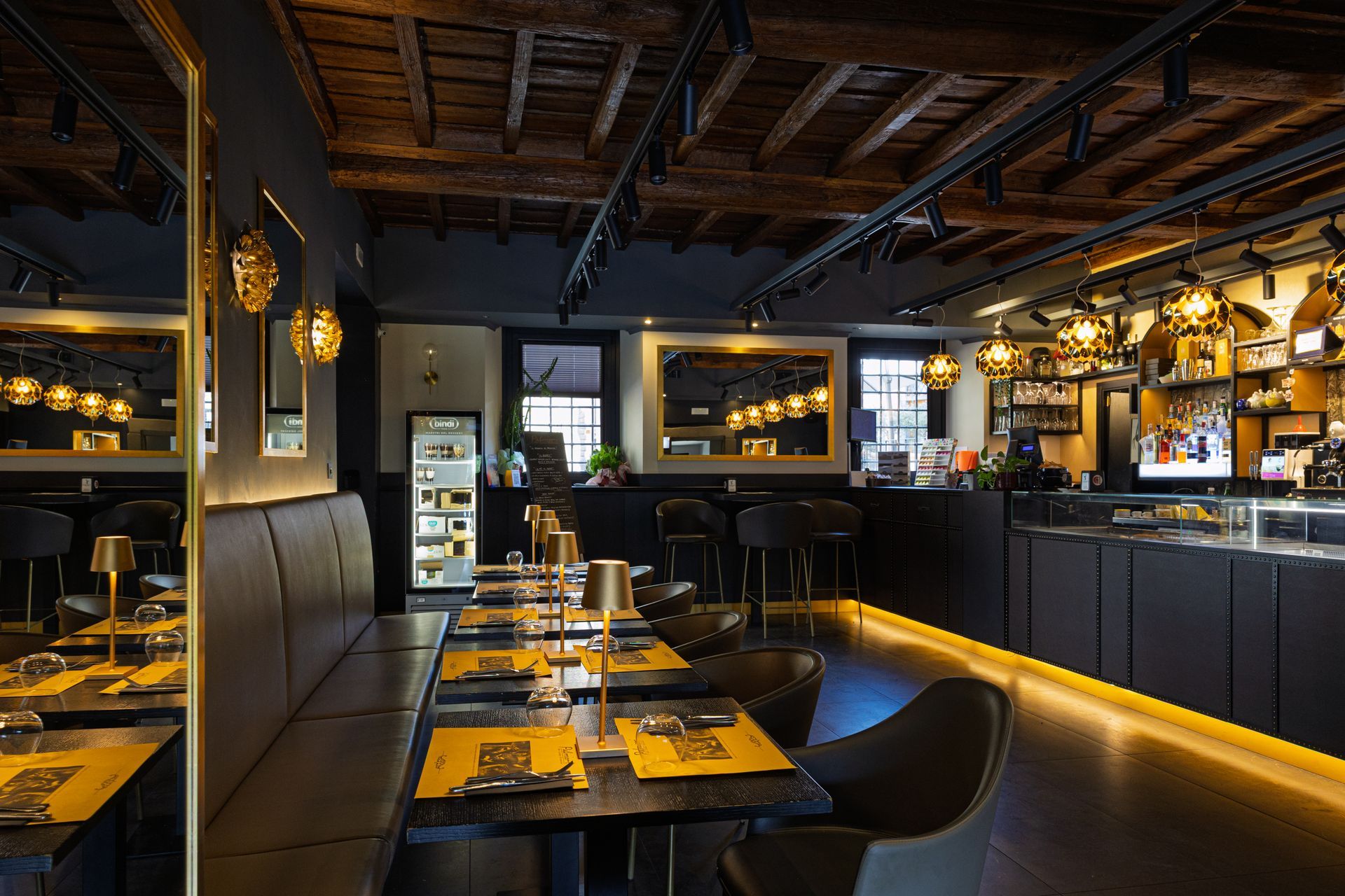 Interno di un ristorante moderno con luci soffuse, tavoli apparecchiati, sedie nere, lampade a sospensione dai toni caldi e un bar sul retro.