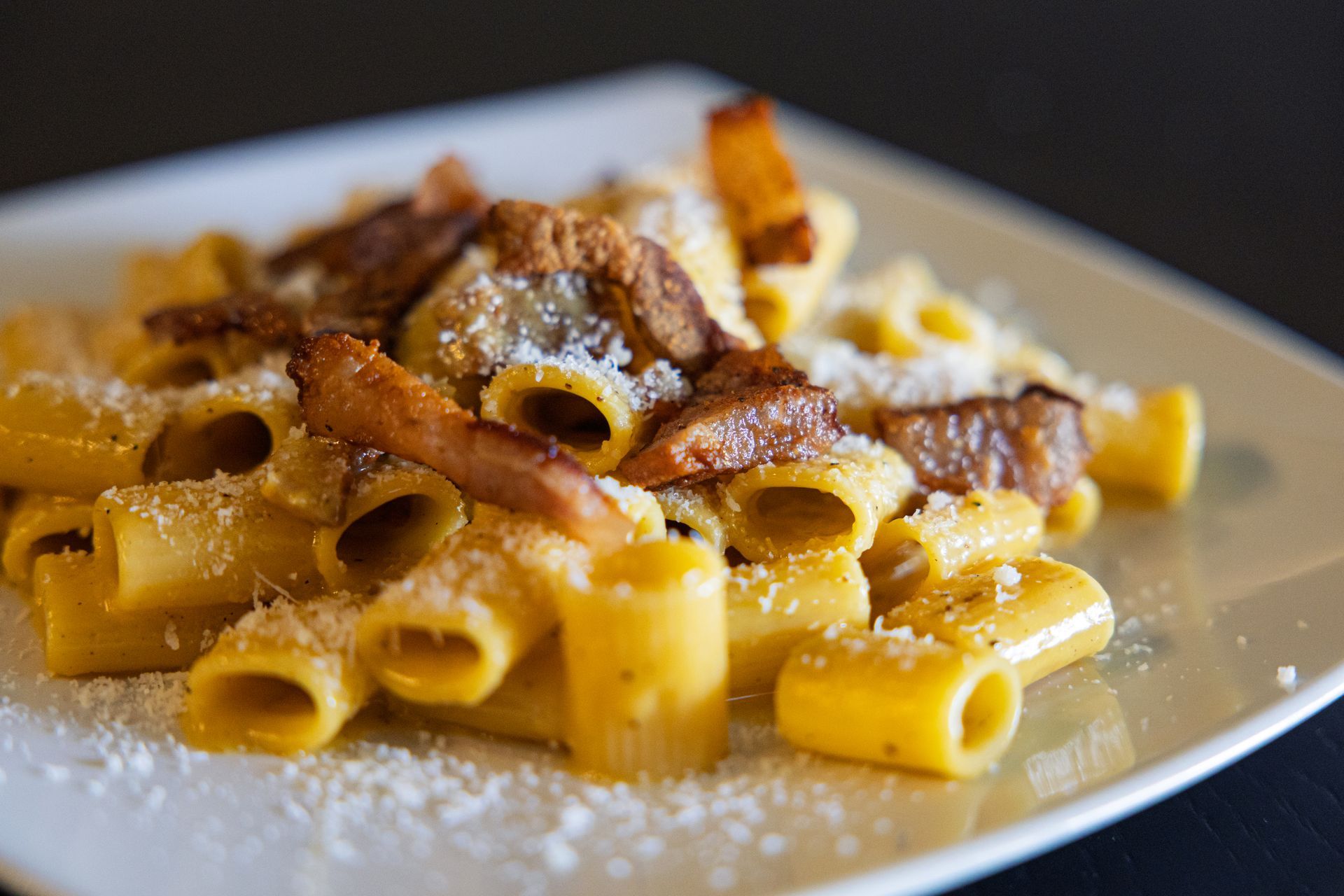 Rigatoni con sugo e pancetta croccante su un piatto bianco, cosparsi di formaggio grattugiato.