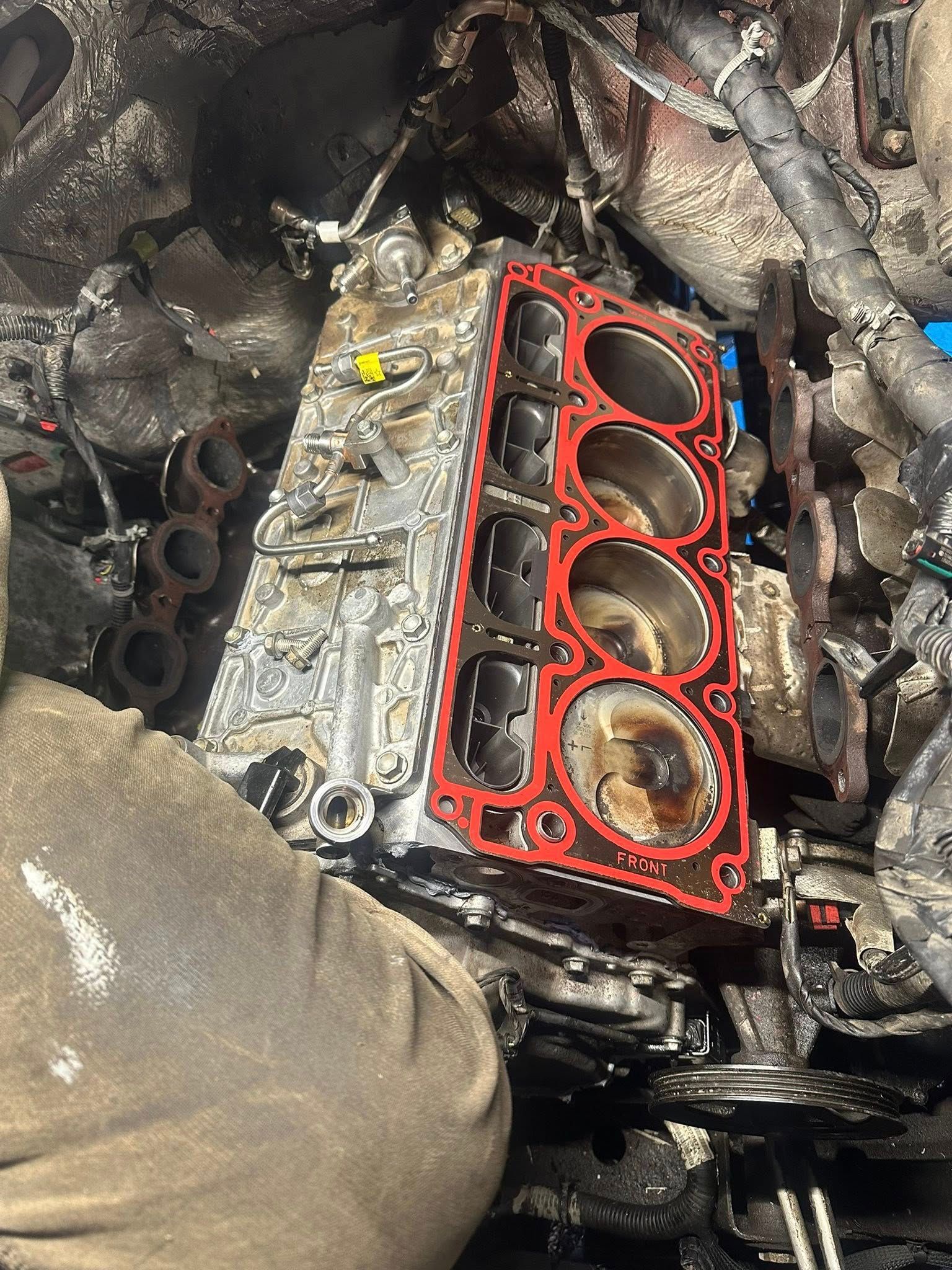 An open engine block with four cylinders, a red gasket, and surrounding mechanical parts in a workshop setting.