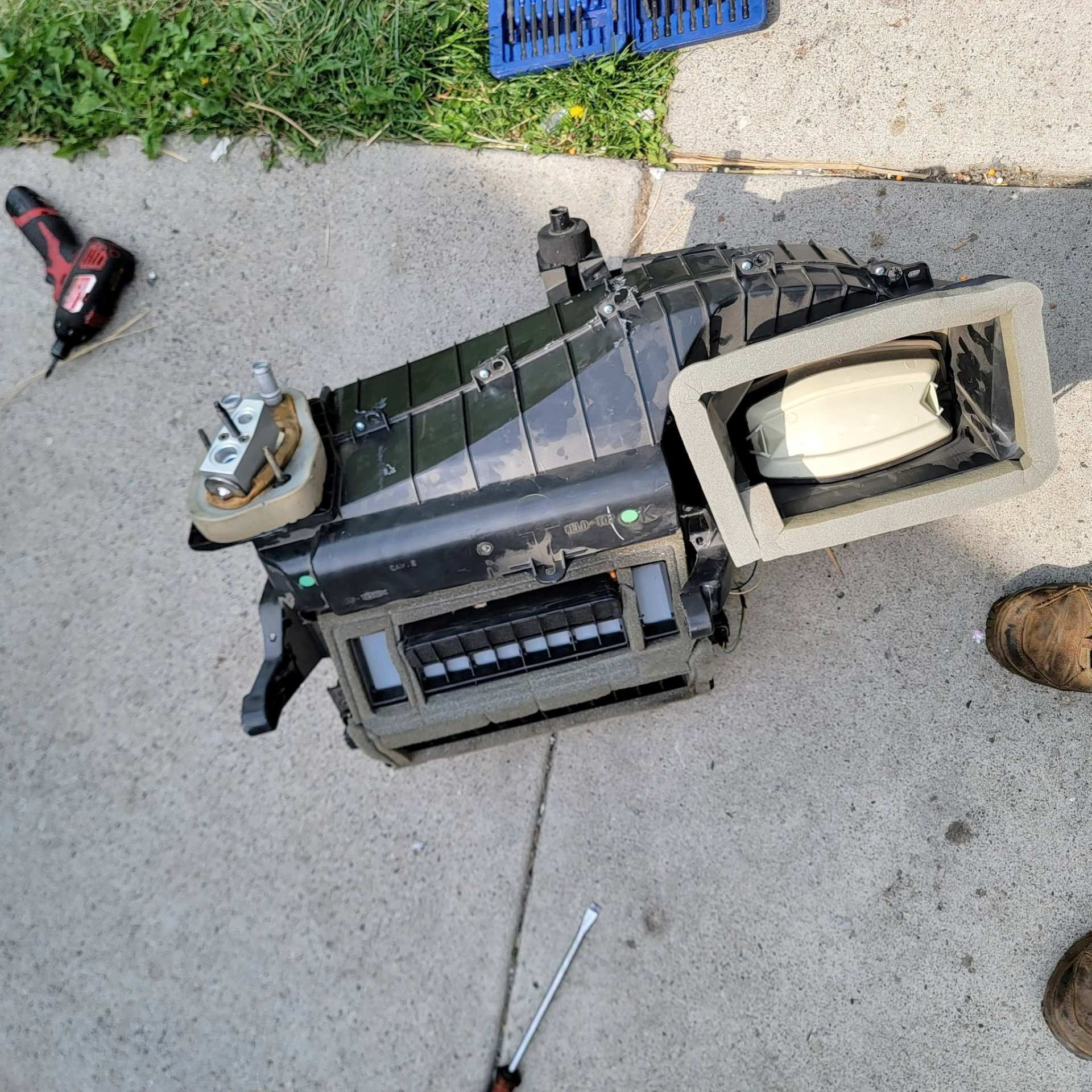 An automotive HVAC blower unit removed from a vehicle, lying on concrete near tools and a shoe.