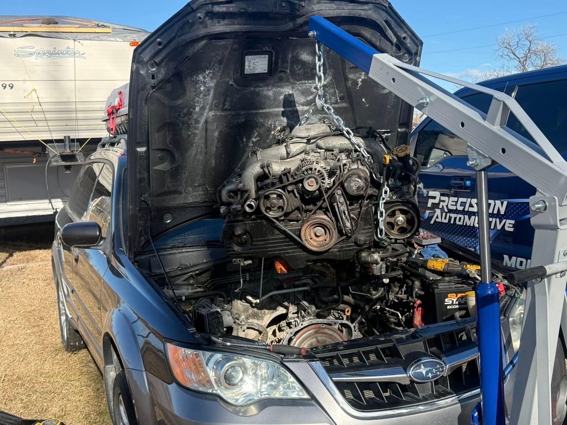An engine hoist suspends a car engine in front of a dark Subaru with its hood open in an outdoor automotive shop area.