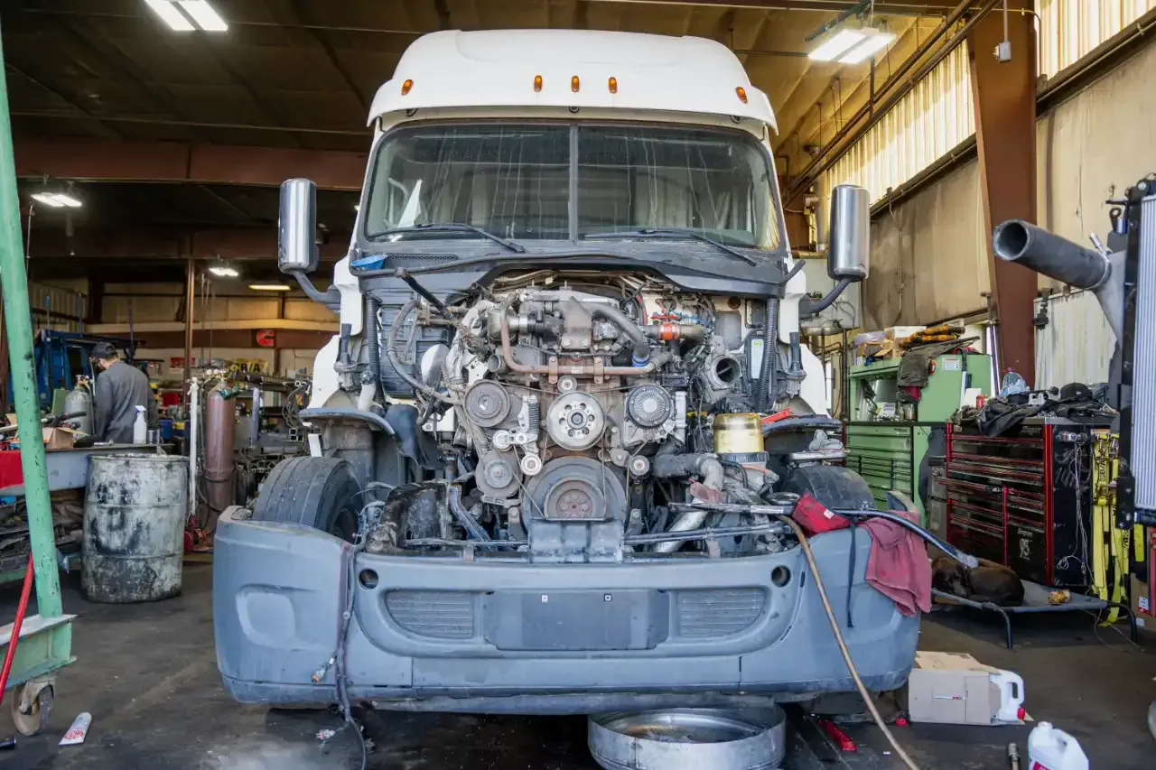 A white semi-truck with its hood open, exposing the engine, sits inside a busy auto repair workshop.
