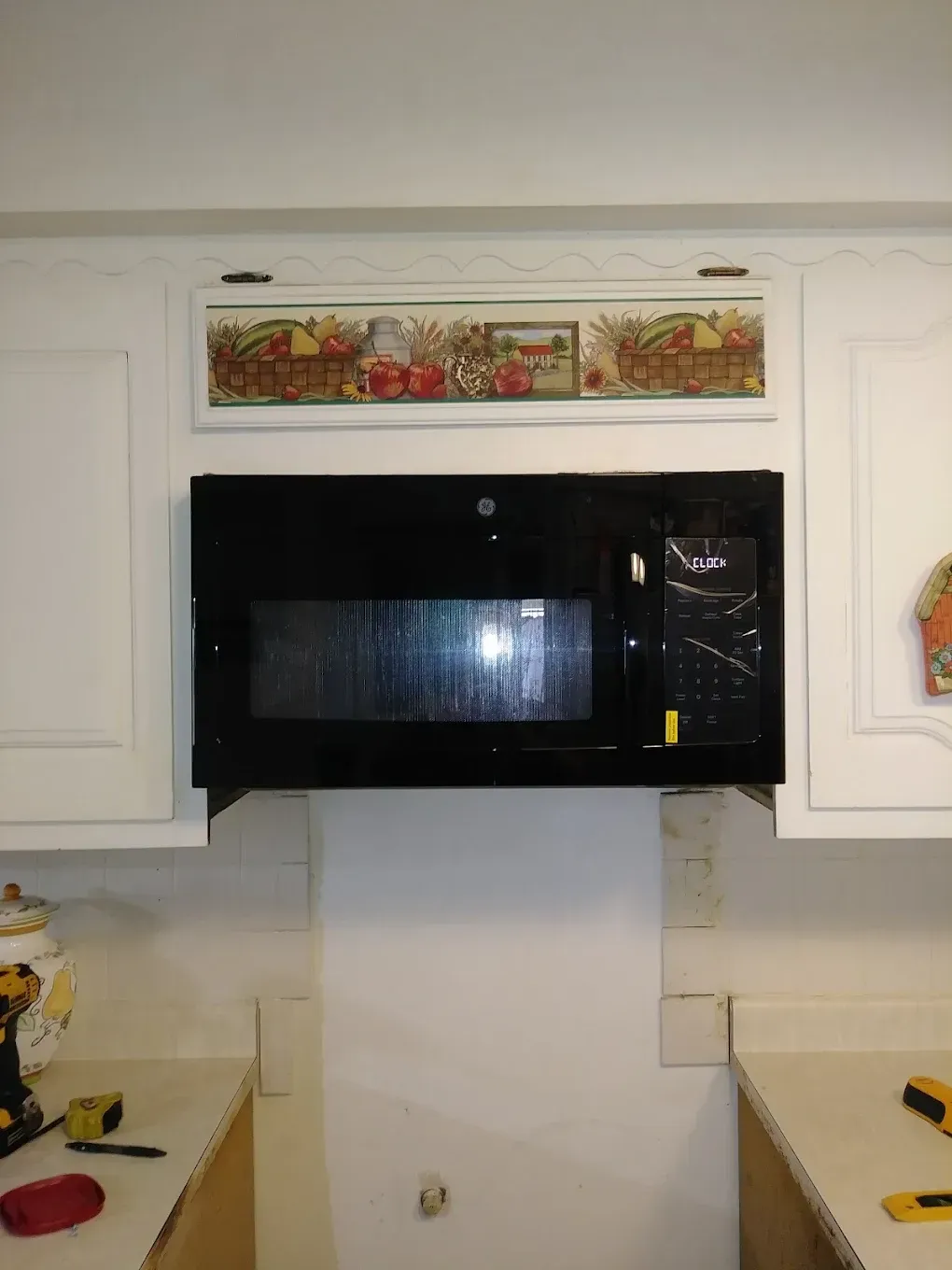 Black microwave mounted between white cabinets, with a decorative border above.