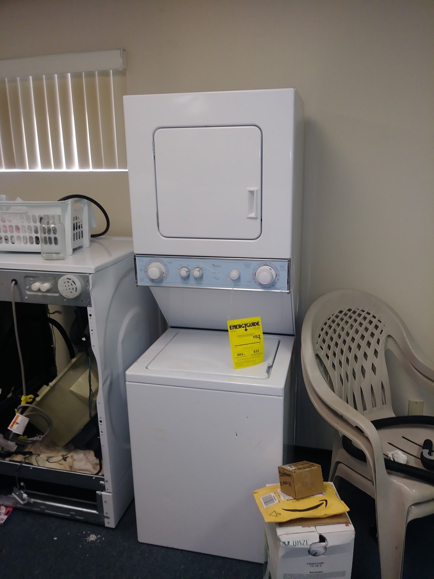 Stackable white washer and dryer with a plastic chair, cardboard box, and another appliance in a room.