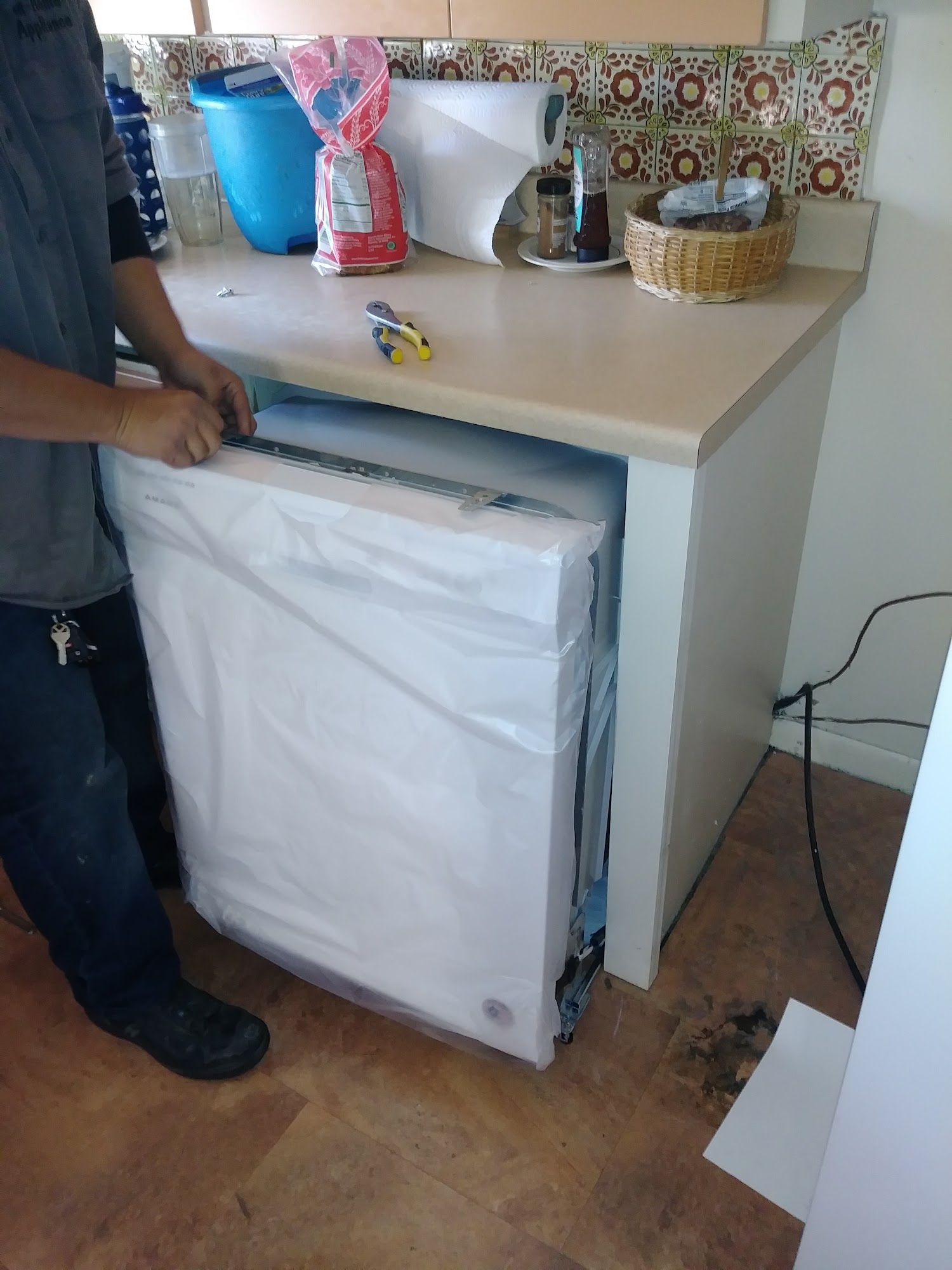 Person installing a dishwasher in a kitchen cabinet. The appliance is wrapped in plastic.