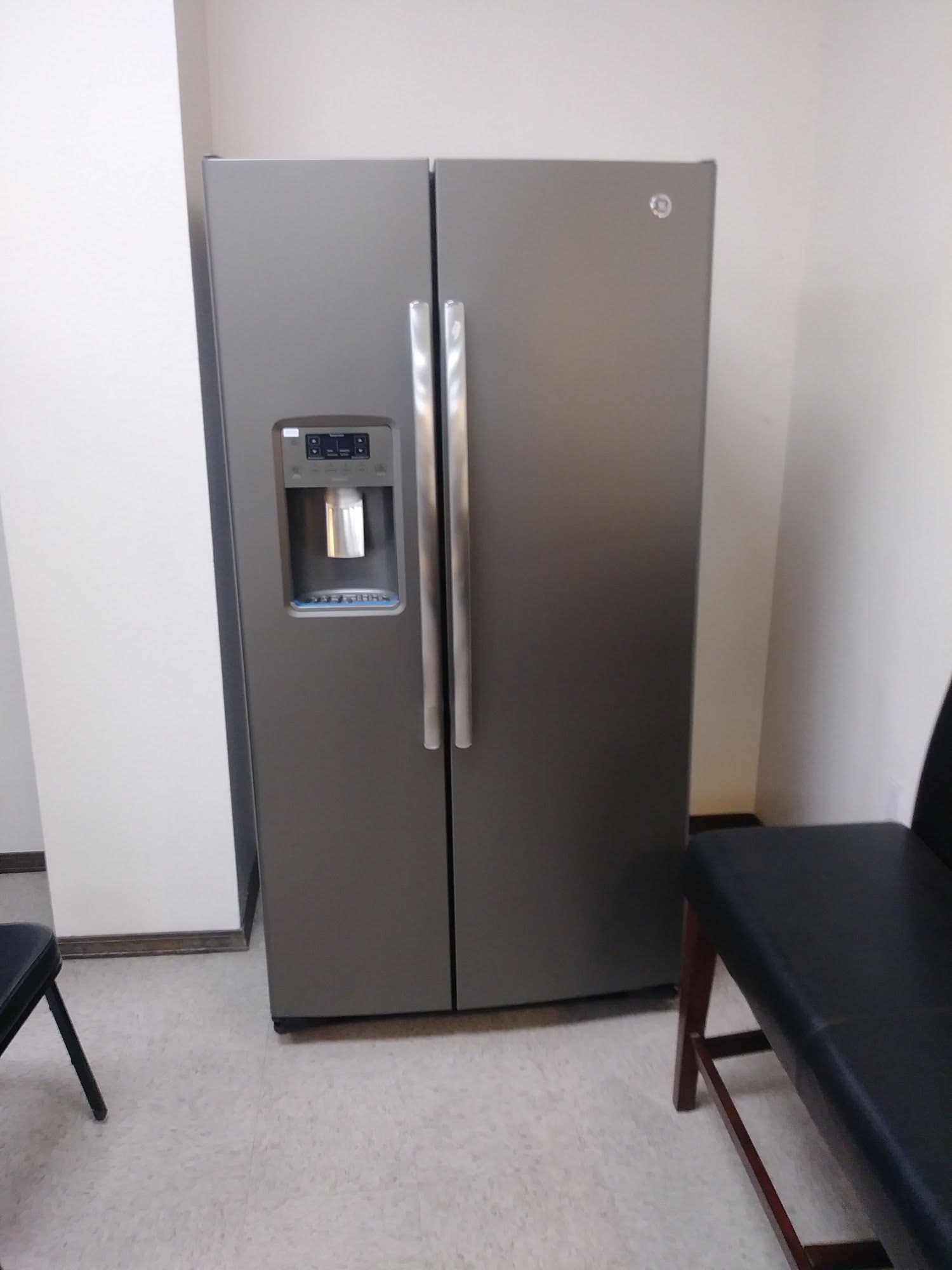 Side-by-side stainless steel refrigerator with water/ice dispenser in a room with carpet, chair and a black bench.