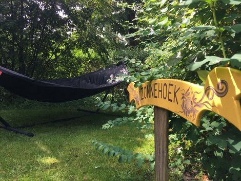 Hangmat in een weelderige tuin naast een geel houten bord en een groene tuinslang.