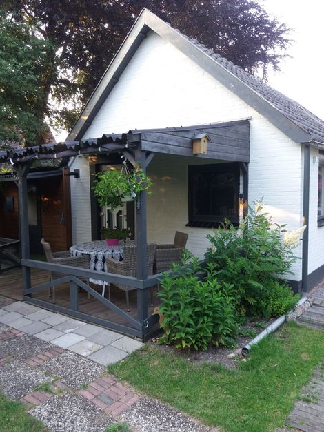 Klein wit huis met een overdekte veranda, zwarte kozijnen, potplanten en een bakstenen pad in een tuin.