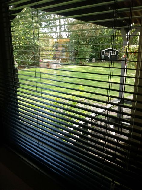 Uitzicht door de donkere jaloezieën op een zonovergoten groene tuin en terras buiten.