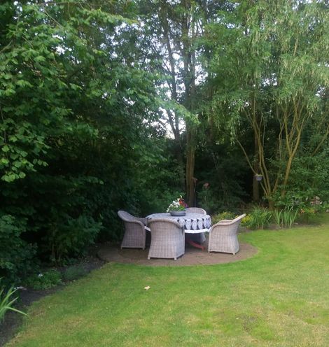 Terrastafel met rieten stoelen op een houten terras in een weelderige groene tuin.