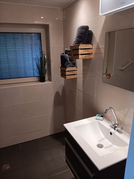 Kleine badkamer met wastafel, spiegel, rolgordijn en opgevouwen handdoeken op wandplanken.