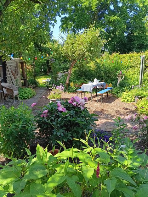 Zonnige tuin met roze bloemen, groene struiken en een picknicktafel onder de bomen.