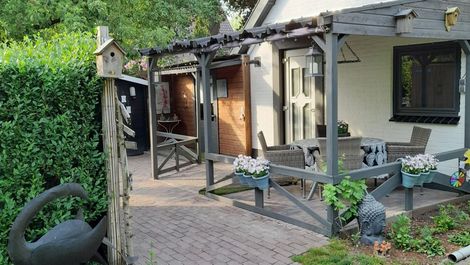 Rustiek tuinterras met met klimplanten begroeide pergola, houten vlonder, stoelen en een decoratieve zwarte zwaan in een plantenbak.