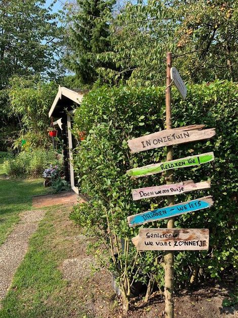 Tuinbordje met kleurrijke pijlen naast een huisje en pad in een zonnige, groene tuin.