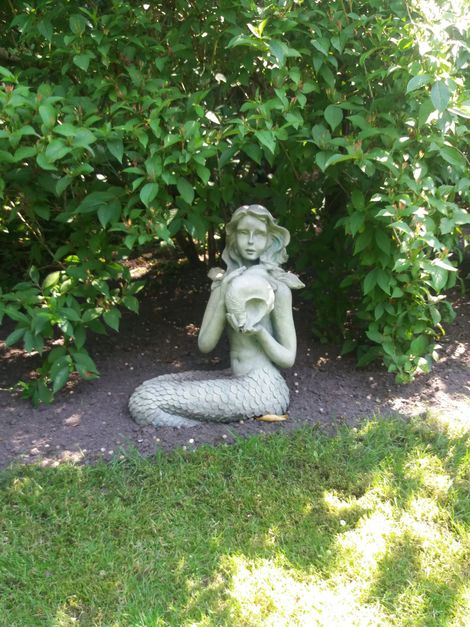 Klein groen zeemeerminbeeldje met een schelp in haar hand, zittend in een tuin tussen bladerrijke struiken en gras.