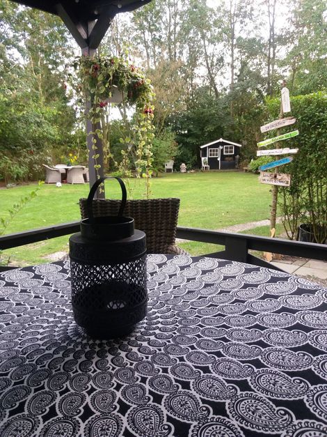 Zwarte lantaarn op een terrastafel met patroon, met uitzicht op een groene achtertuin en tuindecoratie.