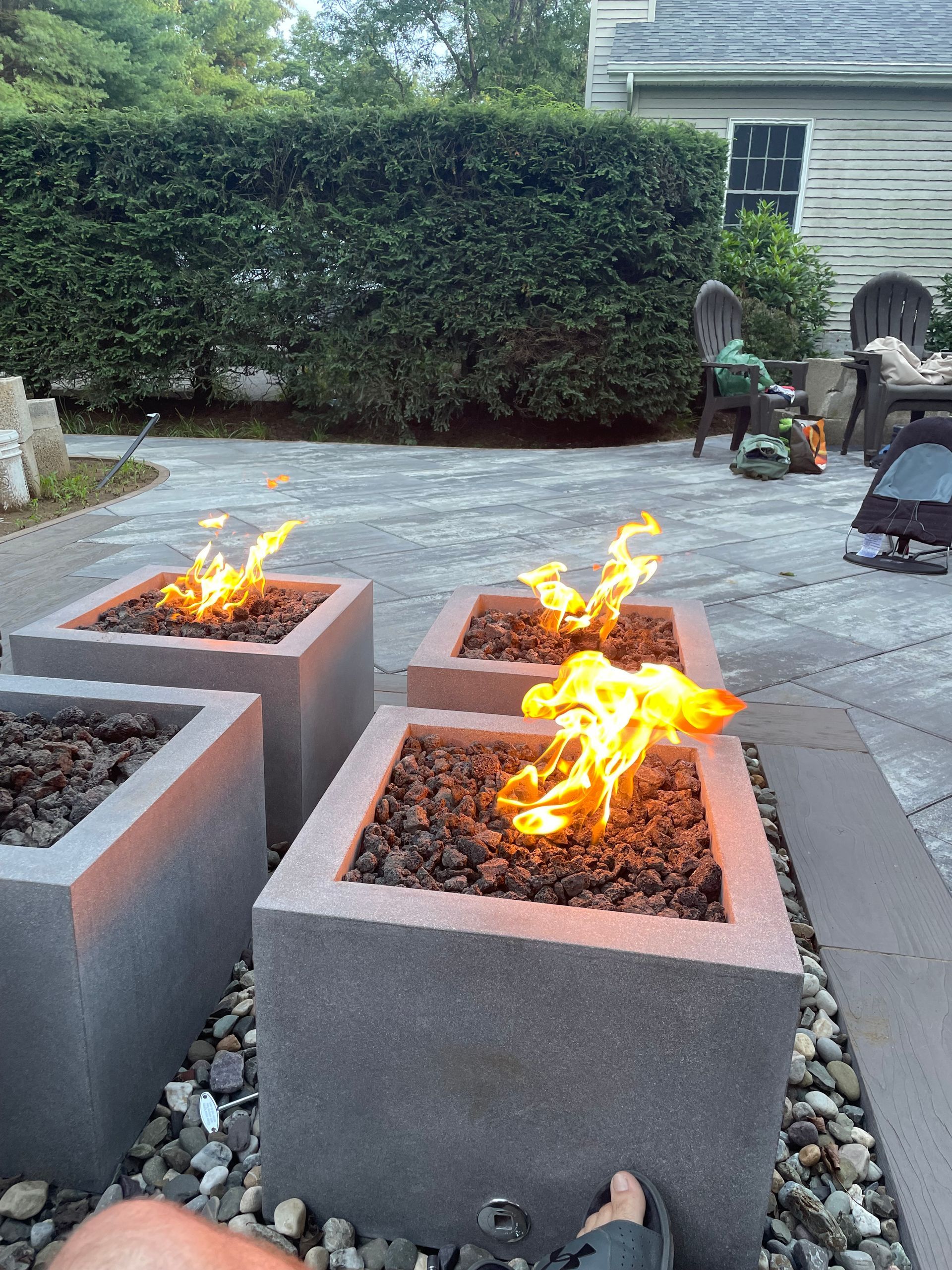 Four square fire pits with flames, set on a stone patio, near a hedge and home.