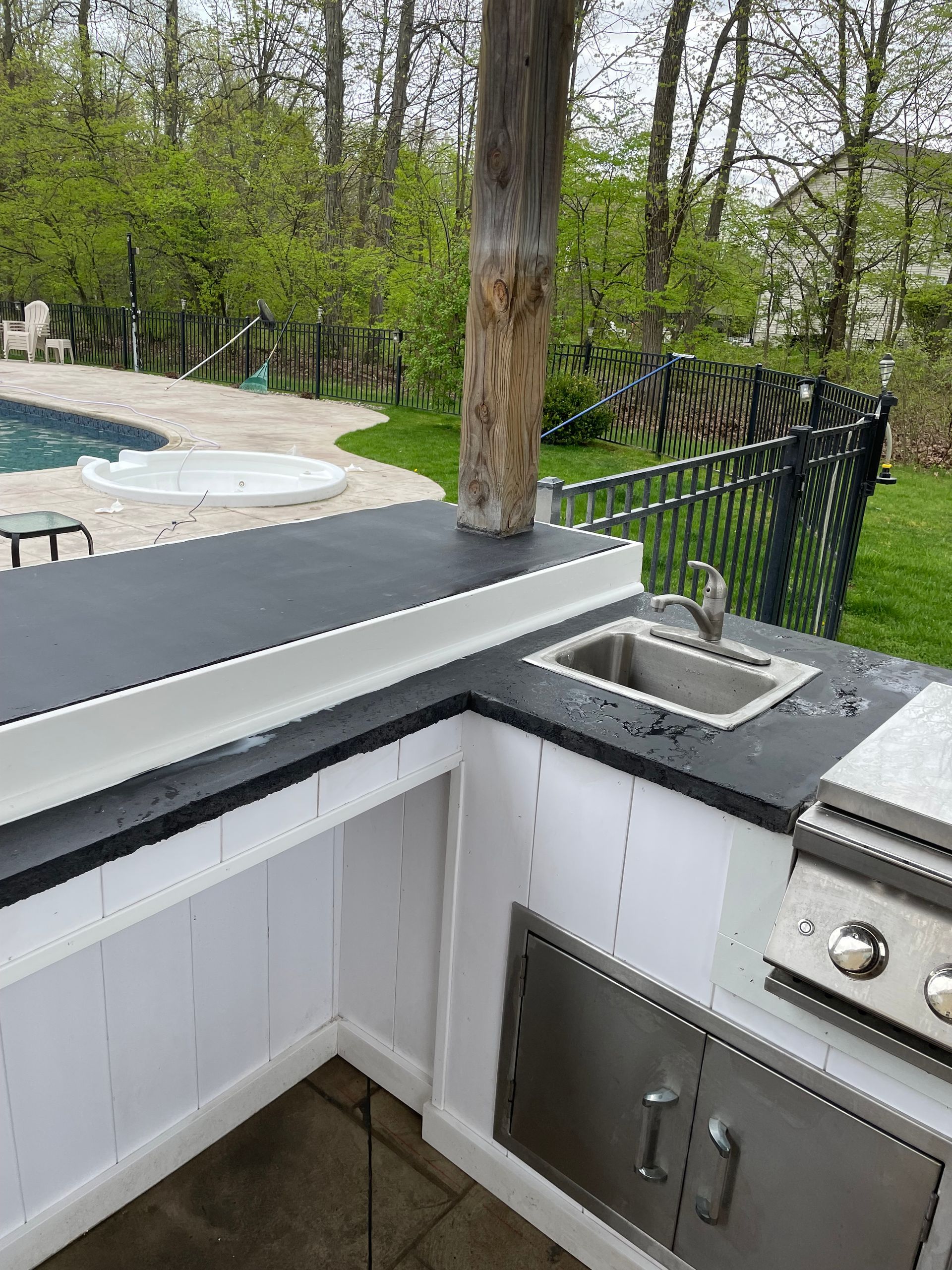 Outdoor kitchen with a sink, grill, and black countertop, set against a pool and green backyard.