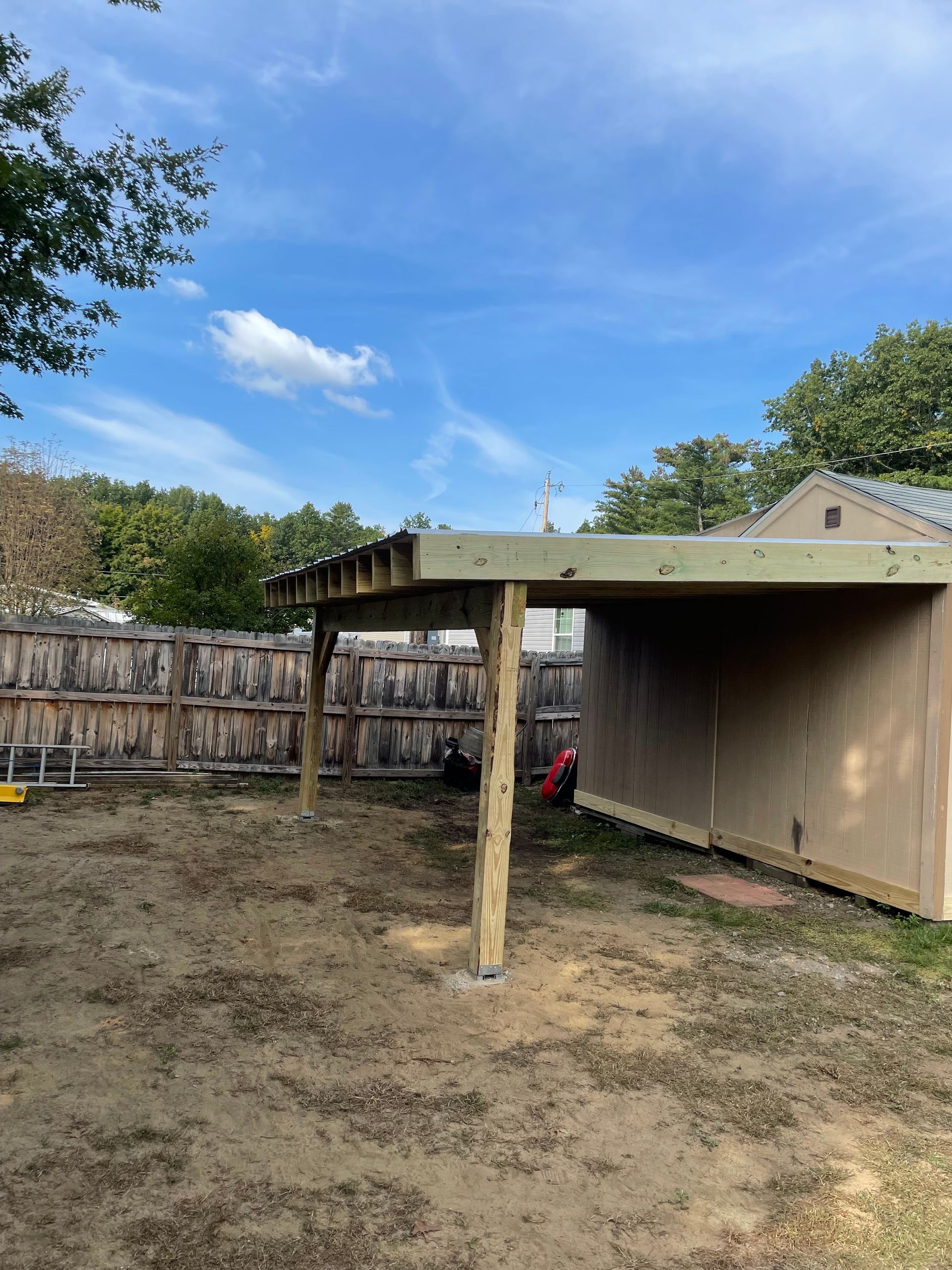 Open-air wooden structure with a roof next to a shed in a yard. Sunny day with blue sky and fence.