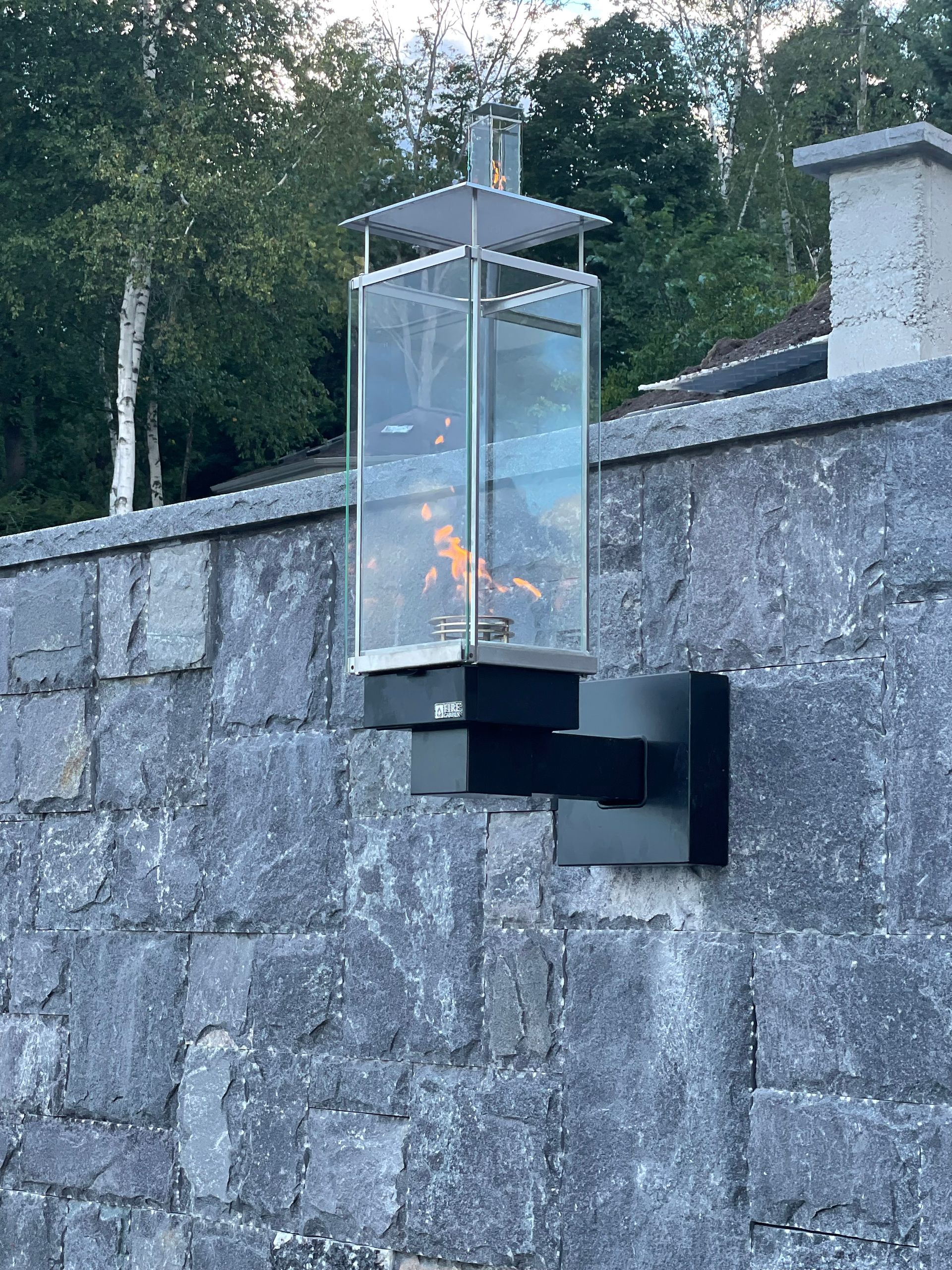 Outdoor gas lantern mounted on a stone wall, flames visible within the glass enclosure.