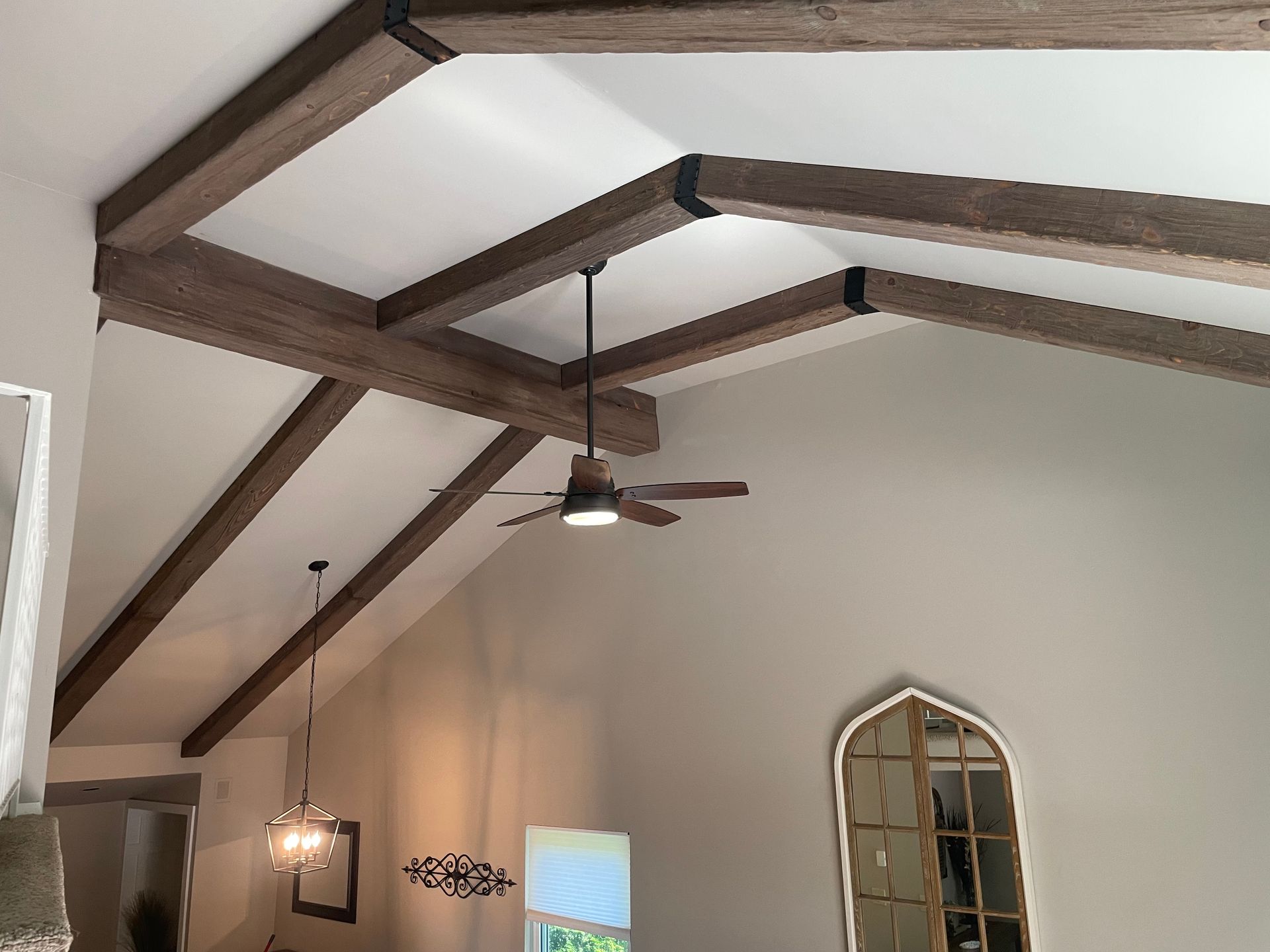 Beamed ceiling with a ceiling fan, a chandelier, and a decorative mirror on the wall.