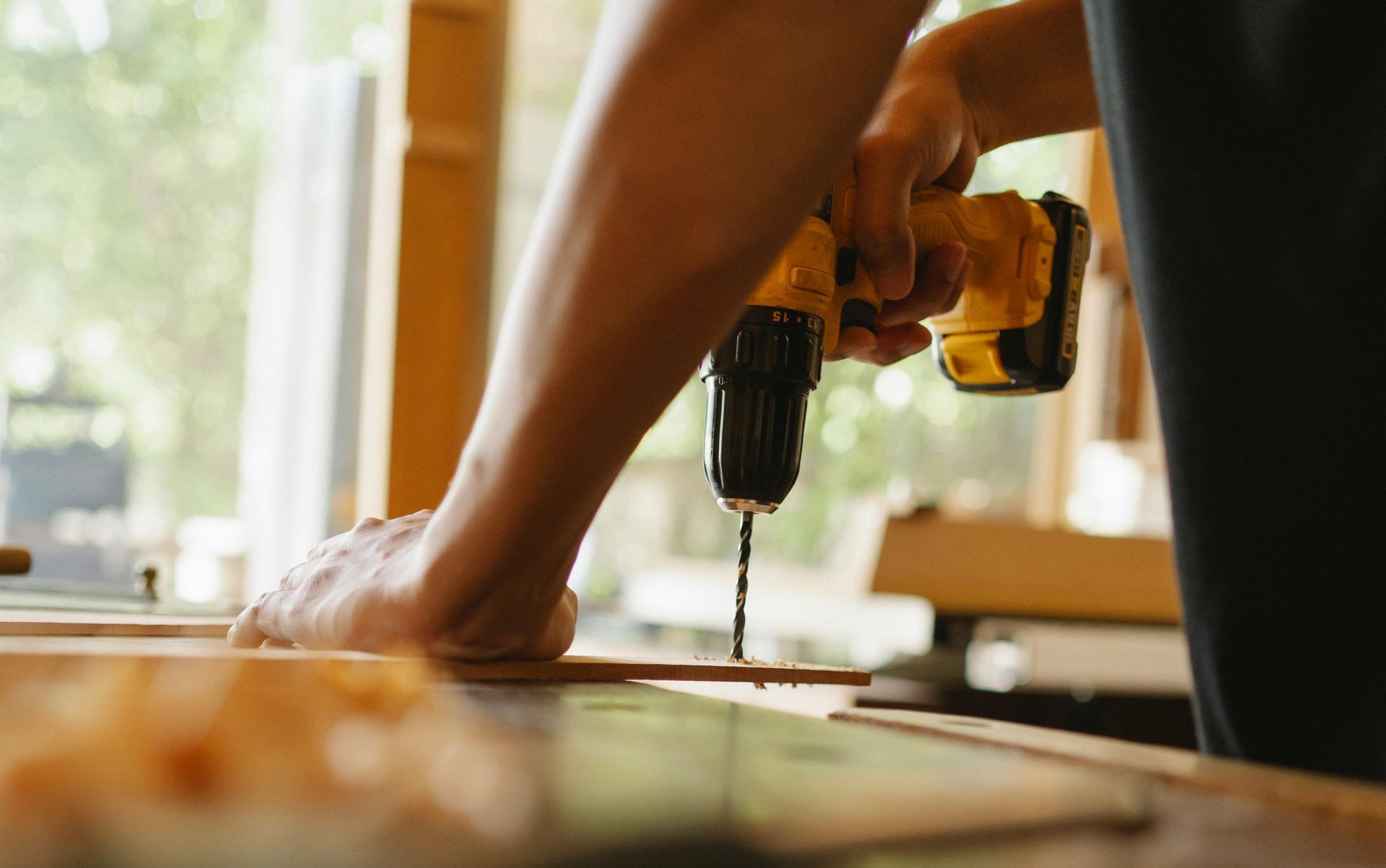 Person using a yellow power drill on a piece of wood indoors, near a window.