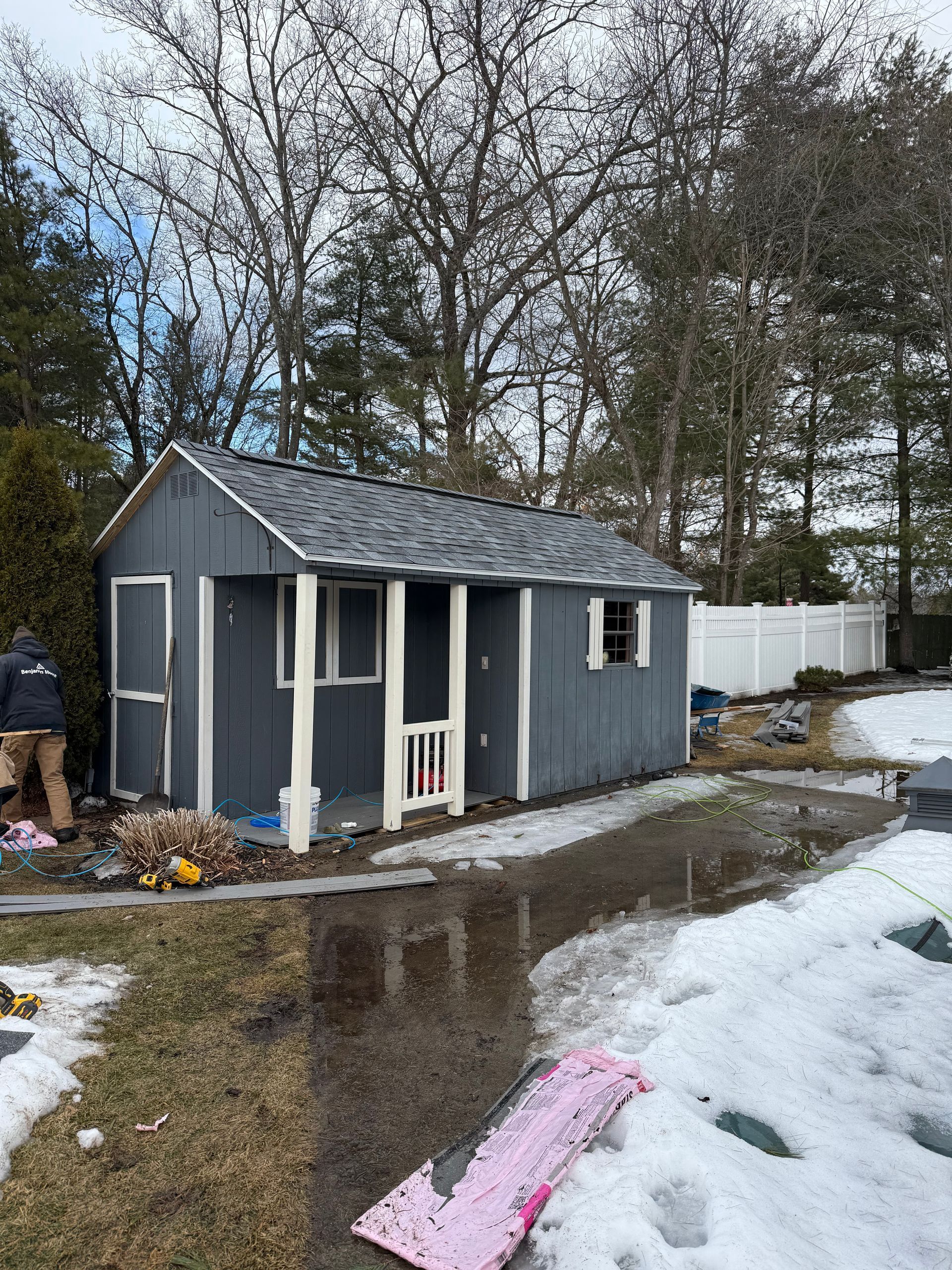 Small gray shed with white trim, in a yard with melting snow, trees, and a white fence.