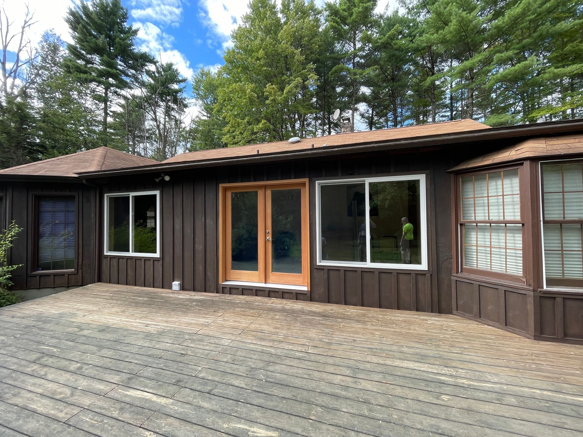 Brown house with windows and doors, wooden deck in front, trees in background.
