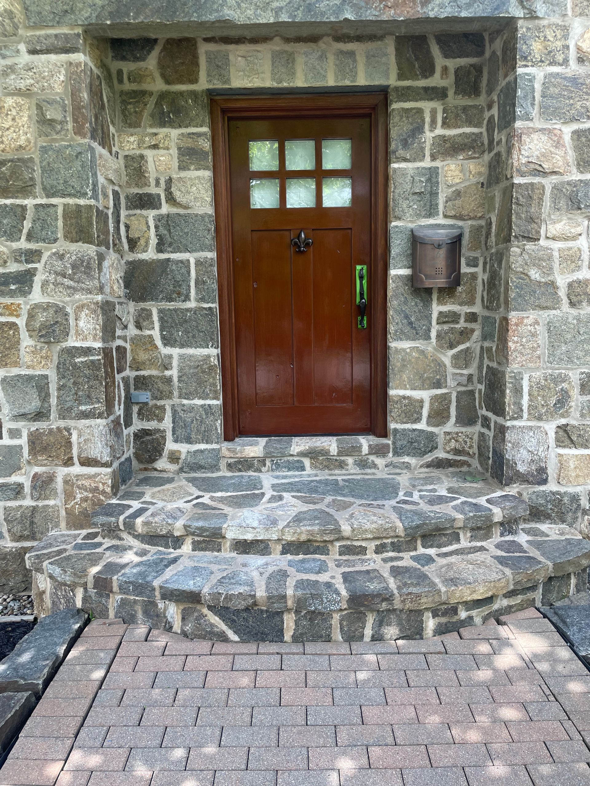 Brown door with grid window set in a stone facade, approached by stone steps and a brick walkway.