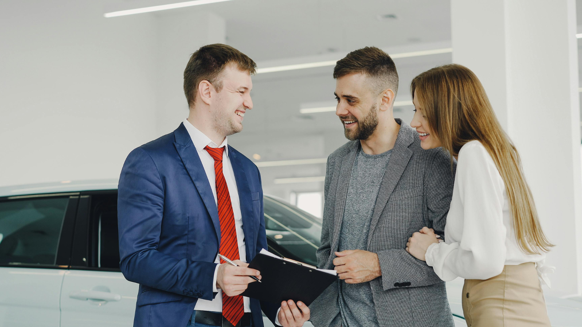 Three people talking beside a car in a bright showroom, one holding a clipboard.