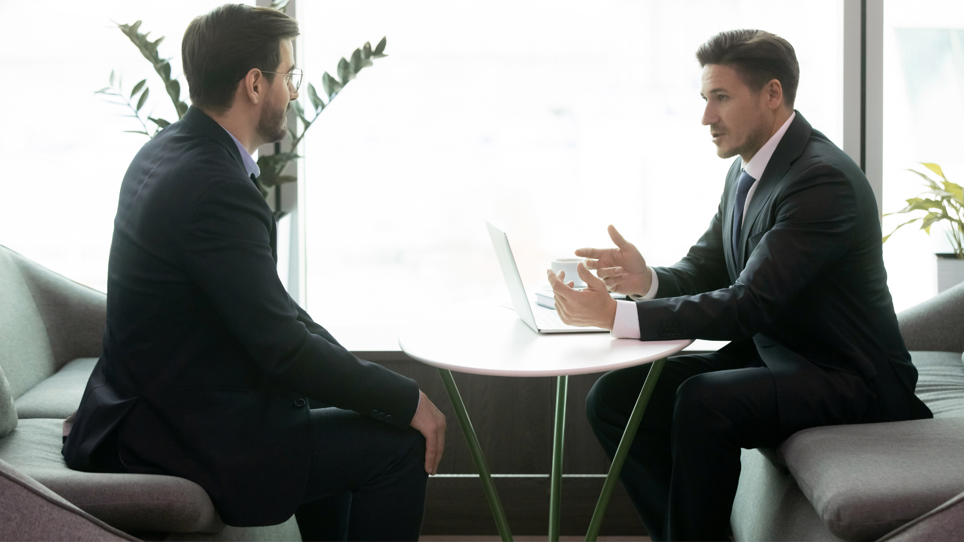Two businessmen in suits discussing across a table with a laptop in a bright office lounge
